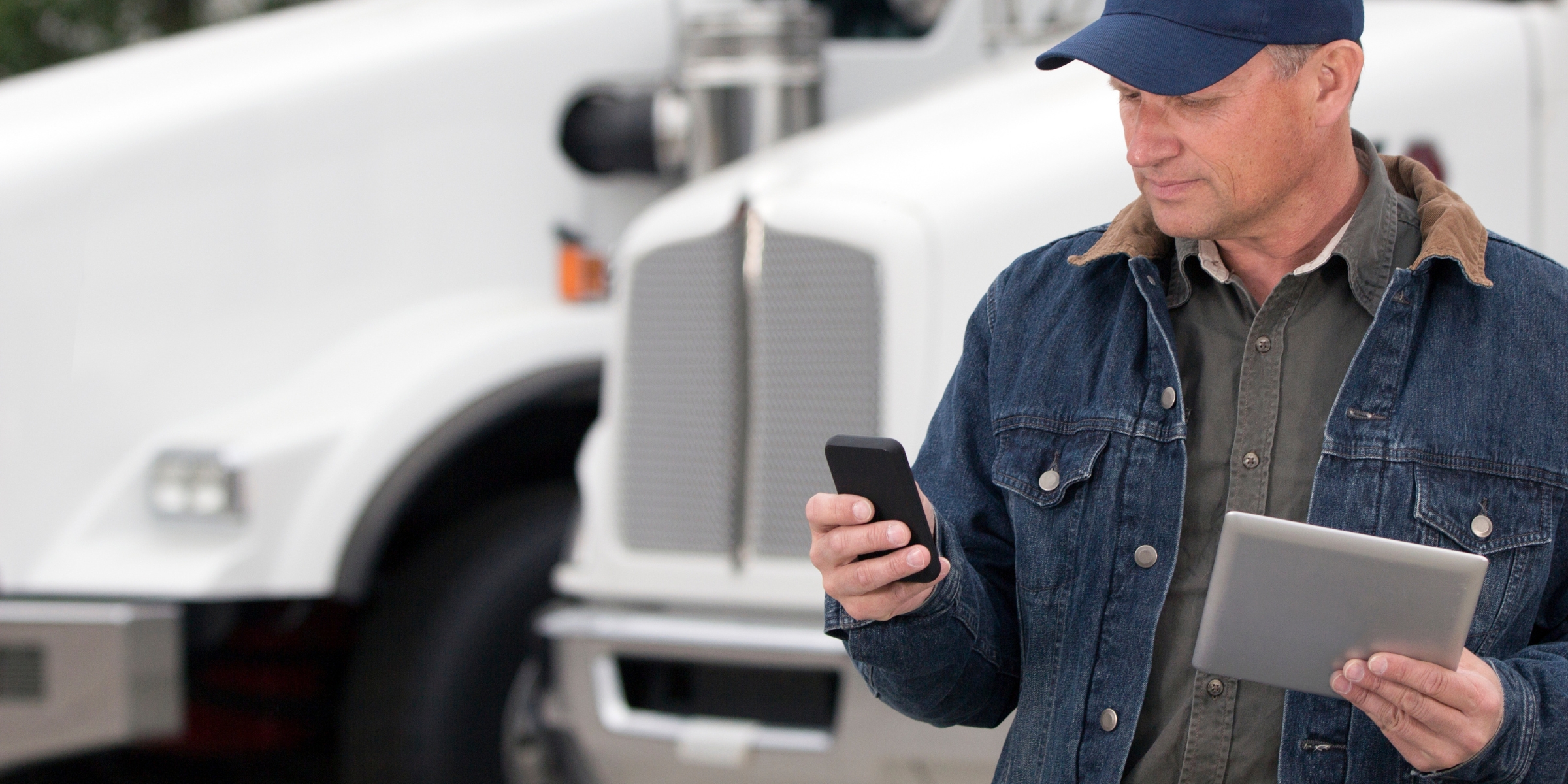 a man wearing a baseball cap and a jean jacket looking at a cell phone as it stands outside in front of a large truck