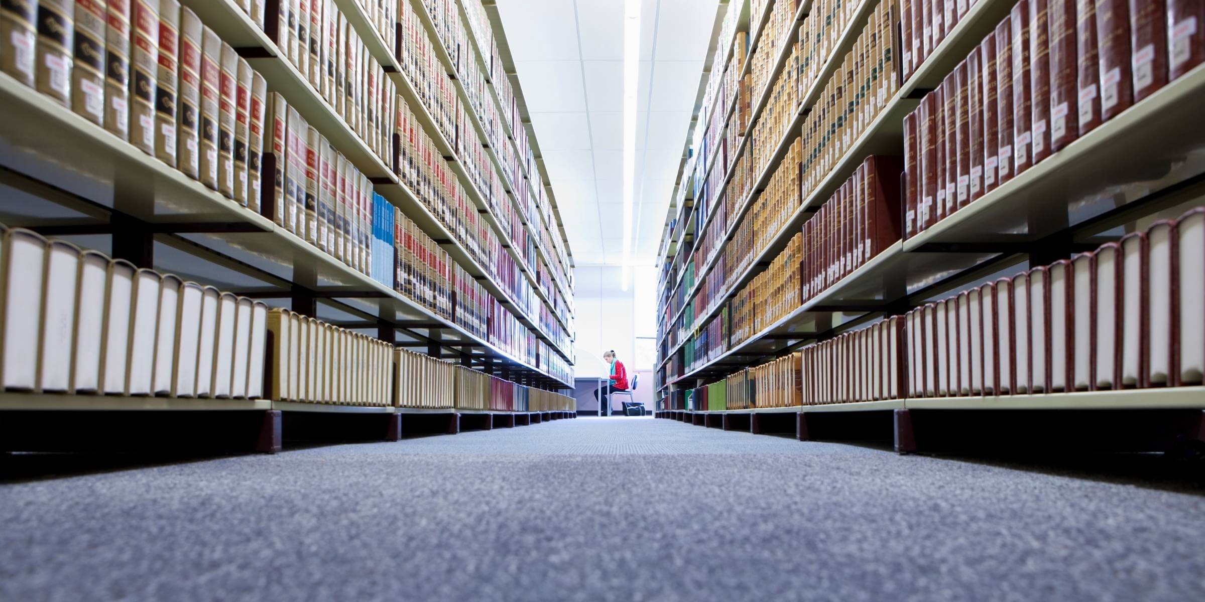 A student sitting and working at a library, surrounded by shelves of books. 