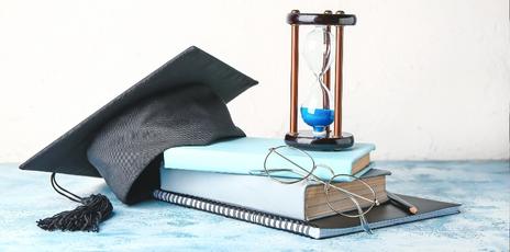 a book sitting on a table next to an hourglass filled with sand, a pair of glasses, and a black grad cap