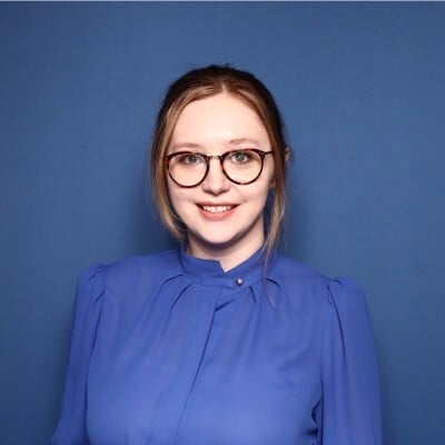 Shauna Cox, Editor-in-Chief of The EvoLLLution, wearing a blue blouse and glasses, smiling against a solid blue backdrop, representing her role in enhancing conversations about non-traditional higher education.