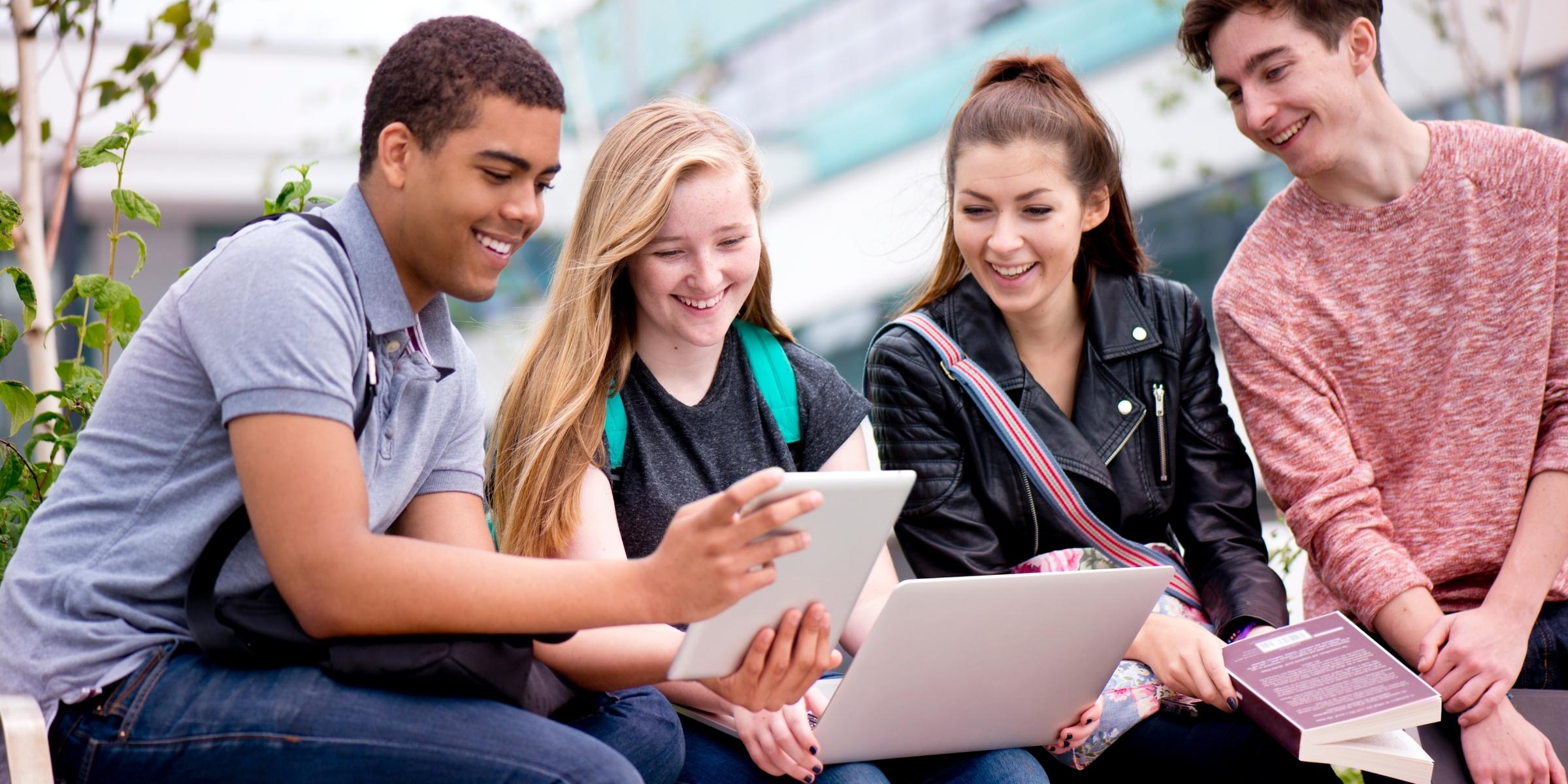 four students sitting outdoors, smiling at mobile devices 